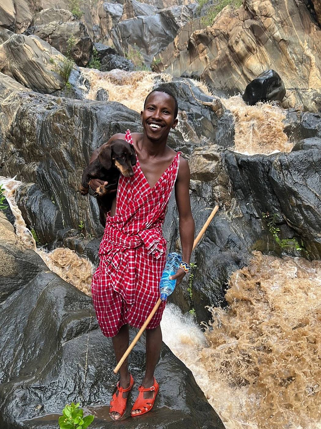 Maasai guide in traditional shuka carrying a dog at the base of a waterfall